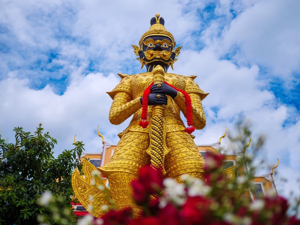 รูปปั้นทองคำขนาดใหญ่ของตัวละครในตำนานตั้งอยู่นอกอาคาร ท่ามกลางท้องฟ้าที่มีเมฆบางส่วนปกคลุม โดยมีดอกไม้สีแดงและ ที่เที่ยวใกล้กรุงเทพ ต้นไม้สีเขียวอยู่ในฉากหน้า—เหมาะสำหรับผู้ที่ต้องการที่เที่ยวใกล้กรุงเทพเพื่อเพลิดเพลินกับศิลปะและธรรมชาติไปพร้อม ๆ กัน.