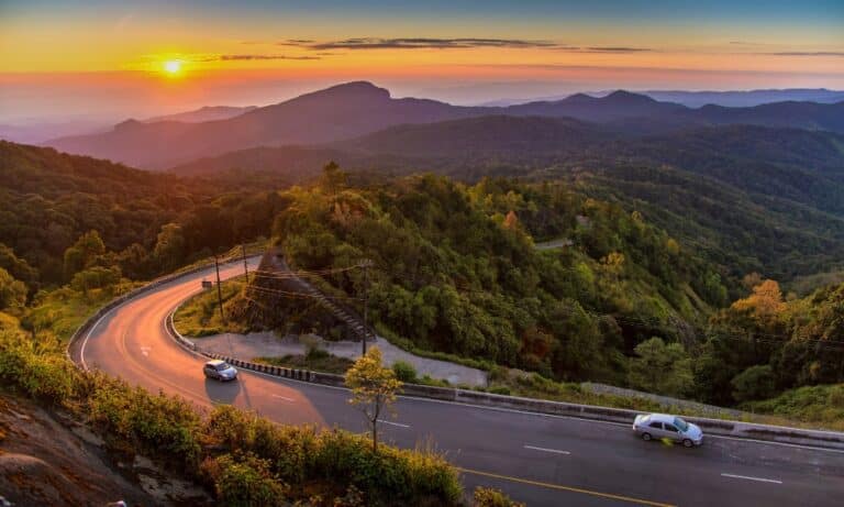 ถนนภูเขาโค้งสองเลนที่มีรถสองคัน ขนาบข้างด้วยป่าเขียวชอุ่มในยามพระอาทิตย์ตกดิน พร้อมด้วยเนินเขาและท้องฟ้าสีสันสดใสเป็นฉากหลัง—ถ่ายทอดความงดงามของทิวทัศน์ ม่อนแจ่มที่เที่ยว ได้อย่างสมบูรณ์แบบ.