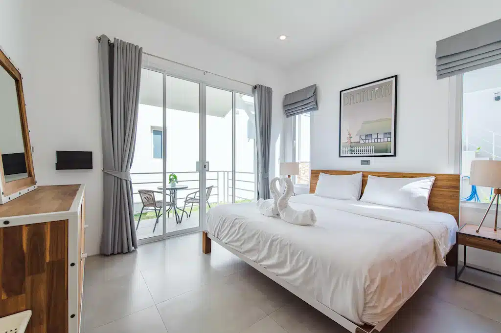 ห้องนอนสไตล์โมเดิร์นที่รีสอร์ทปราณบุรี พร้อมเตียงขนาดใหญ่ ผ้าปูที่นอนสีขาว ผ้าเช็ดตัวรูปหงส์ เฟอร์นิเจอร์ไม้ หน้าต่างบานใหญ่พร้อมผ้าม่านสีเทา และระเบียงส่วนตัว.