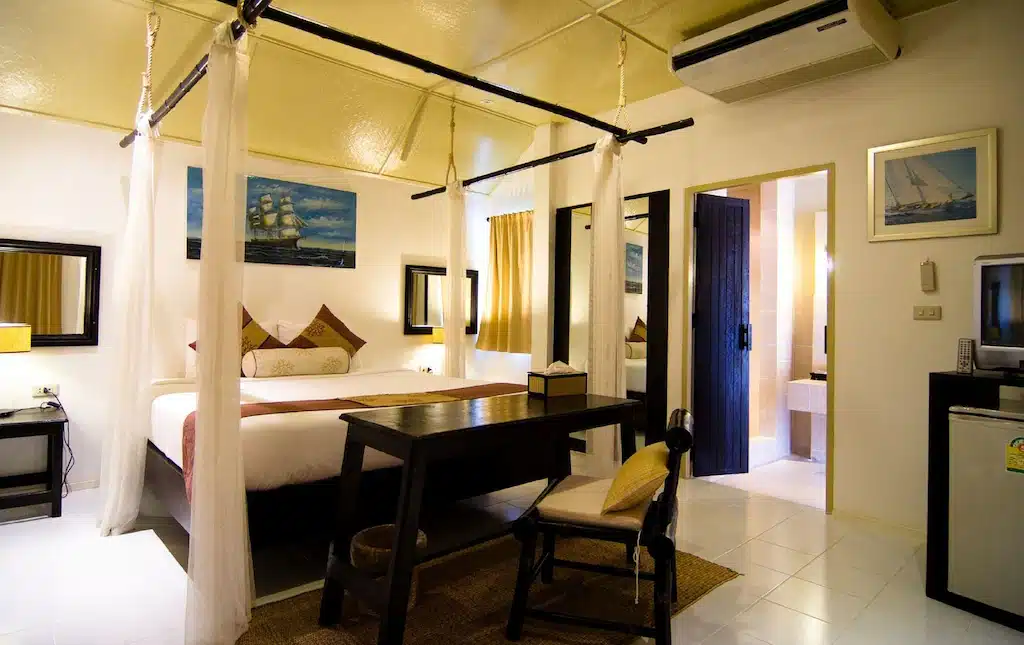 ห้องพักโรงแรม ปราณบุรี ที่จัดเรียงอย่างเรียบร้อย พร้อมเตียงมีหลังคาคลุม โต๊ะทำงาน เก้าอี้ เครื่องปรับอากาศ งานศิลปะบนผนัง ตู้เย็น และประตูห้องน้ำแบบเปิดโล่ง.