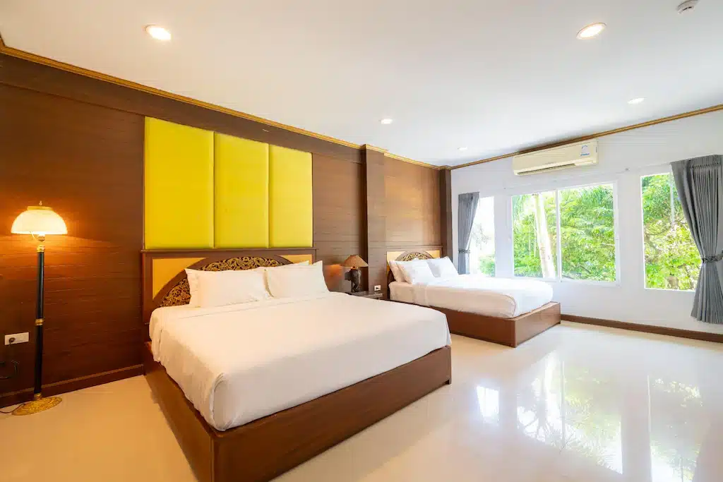 ห้องพักโรงแรมพร้อมเตียงคู่สองเตียง หัวเตียงสีเหลือง ผนังไม้ ไฟตั้งพื้น พื้นกระเบื้องสีขาว หน้าต่างบานใหญ่พร้อมผ้าม่านสีเทา และเครื่องปรับอากาศ—เหมาะสำหรับผู้ที่มองหาที่พักประจวบหรือโรงแรมประจวบที่สะดวกสบาย.