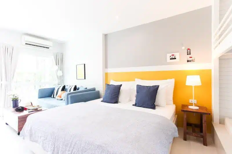 ห้องนอนสว่างสดใสที่รีสอร์ทปราณบุรี พร้อมเตียงคู่ ผ้าปูที่นอนสีน้ำเงินและขาว โต๊ะข้างเตียงพร้อมโคมไฟ โซฟาสีน้ำเงิน หน้าต่างบานใหญ่ และผนังตกแต่งสีสันสดใส—เหมาะสำหรับการพักผ่อนของคุณ.