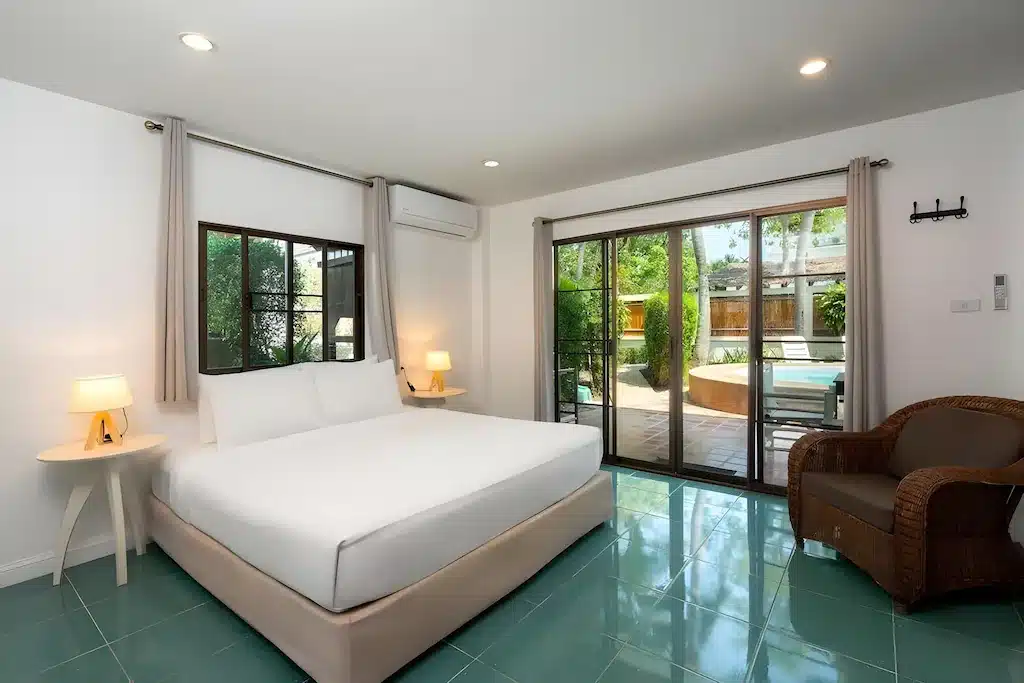 ห้องนอนสมัยใหม่พร้อมเตียงคู่ โต๊ะข้างเตียงสองตัวพร้อมโคมไฟ เก้าอี้หวาย และประตูกระจกที่เปิดออกสู่ระเบียงพร้อมสระว่ายน้ำด้านนอก—เหมาะสำหรับการพักผ่อนที่ ที่พักประจวบติดทะเล.