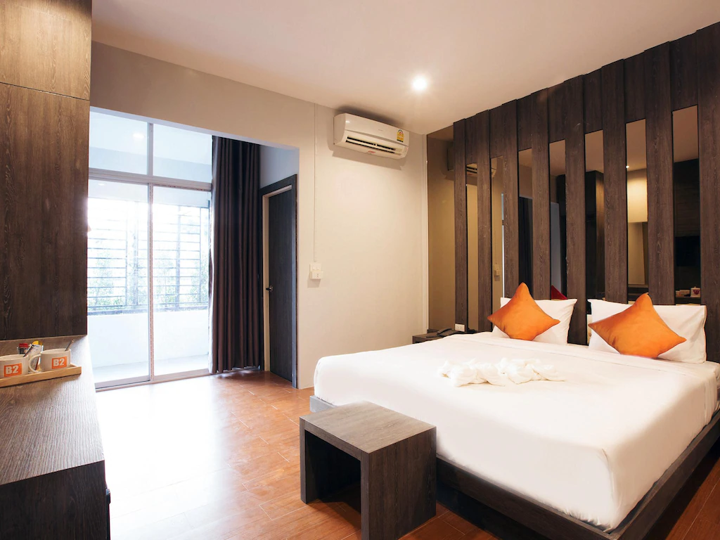 ห้องพักโรงแรมทันสมัยพร้อมเตียงคู่ขนาดใหญ่ หมอนอิงสีส้ม หัวเตียงกระจกเงา เฟอร์นิเจอร์ไม้ เครื่องปรับอากาศ และระเบียง—เหมาะสำหรับการพักผ่อนหลังจากสำรวจสถานที่ท่องเที่ยวในบุรีรัมย์และสถานที่ท่องเที่ยวอื่นๆ ในบุรีรัมย์ที่อยู่ใกล้เคียง.