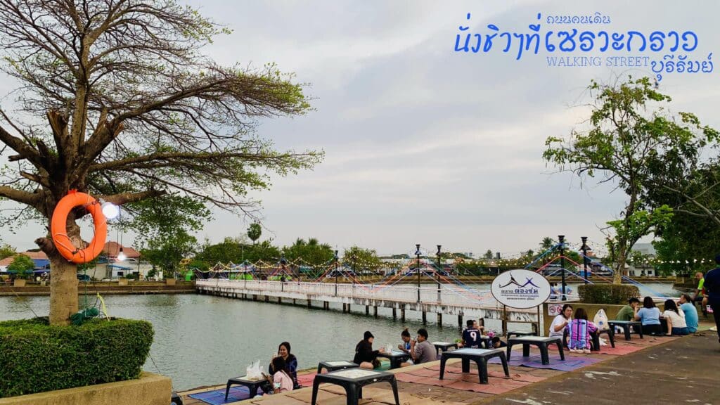 ผู้คนนั่งที่โต๊ะเตี้ยริมทางเดินเลียบแม่น้ำในหนึ่งในสถานที่ท่องเที่ยวที่มีเสน่ห์ของบุรีรัมย์ โดยมีสะพานข้ามแม่น้ำเป็นฉากหลัง ต้นไม้ที่มีเสื้อชูชีพตั้งอยู่ด้านหน้า และมีข้อความภาษาไทยปรากฏอยู่ที่มุมขวาบน.
