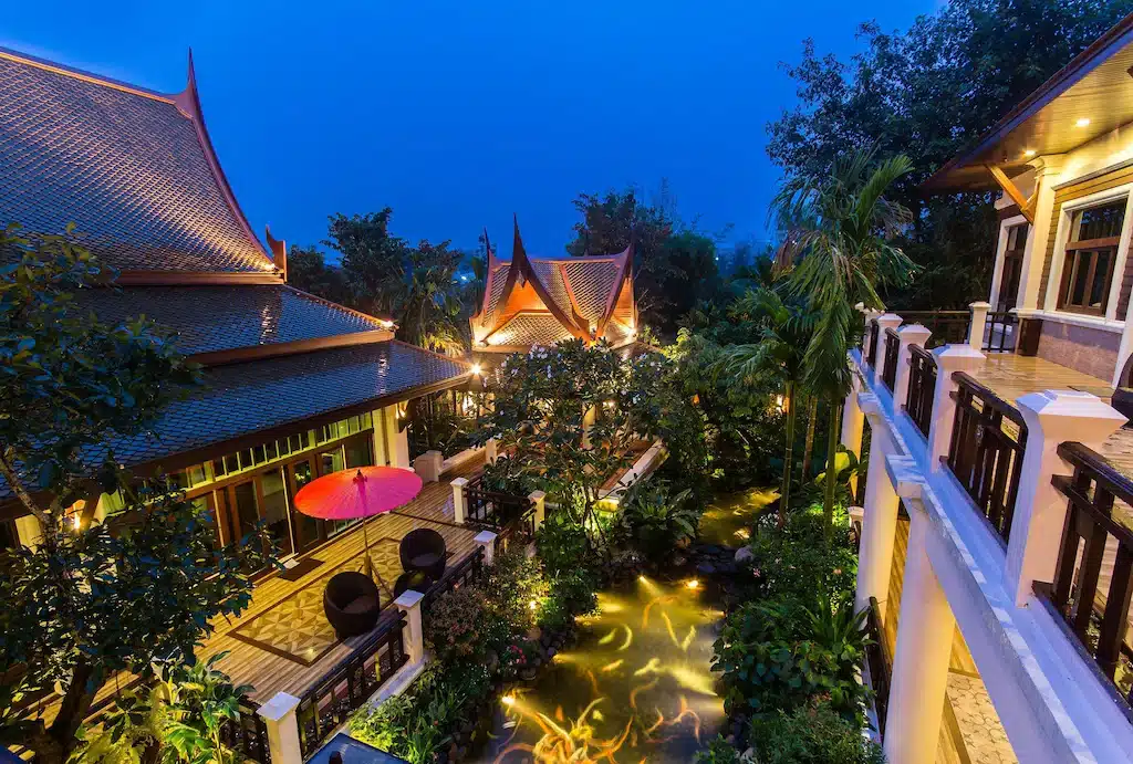 อาคารสไตล์ไทยดั้งเดิมที่มีหลังคาแหลมล้อมรอบลานสวนซึ่งประดับด้วยแสงไฟอบอุ่น มีพืชเขตร้อนและบ่อน้ำเล็กๆ มองเห็นได้ในยามพลบค่ำ—เป็นตัวเลือกที่น่าดึงดูดสำหรับ ที่พักเชียงใหม่ธรรมชาติ.