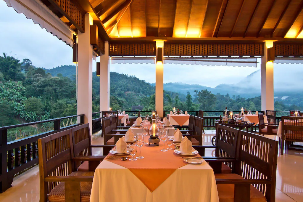 ร้านอาหารกลางแจ้งที่มีโต๊ะและเก้าอี้ไม้จัดเรียงอย่างเรียบร้อย มองเห็นทิวทัศน์ภูเขาเขียวขจีภายใต้ท้องฟ้าที่มีเมฆปกคลุม—เหมาะสำหรับผู้ที่มองหาที่พักเชียงใหม่ท่ามกลางธรรมชาติ.