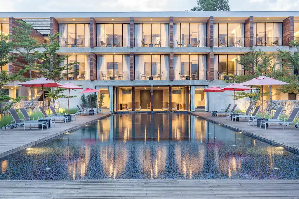 โรงแรมประจวบสไตล์โมเดิร์นพร้อมสองชั้นของระเบียงกระจกที่มองเห็นสระว่ายน้ำกลางแจ้งรูปสี่เหลี่ยมผืนผ้า ล้อมรอบด้วยเก้าอี้อาบแดดและร่มสีแดง—เหมาะอย่างยิ่งสำหรับประสบการณ์การพักผ่อนของคุณที่โรงแรมประจวบ.
