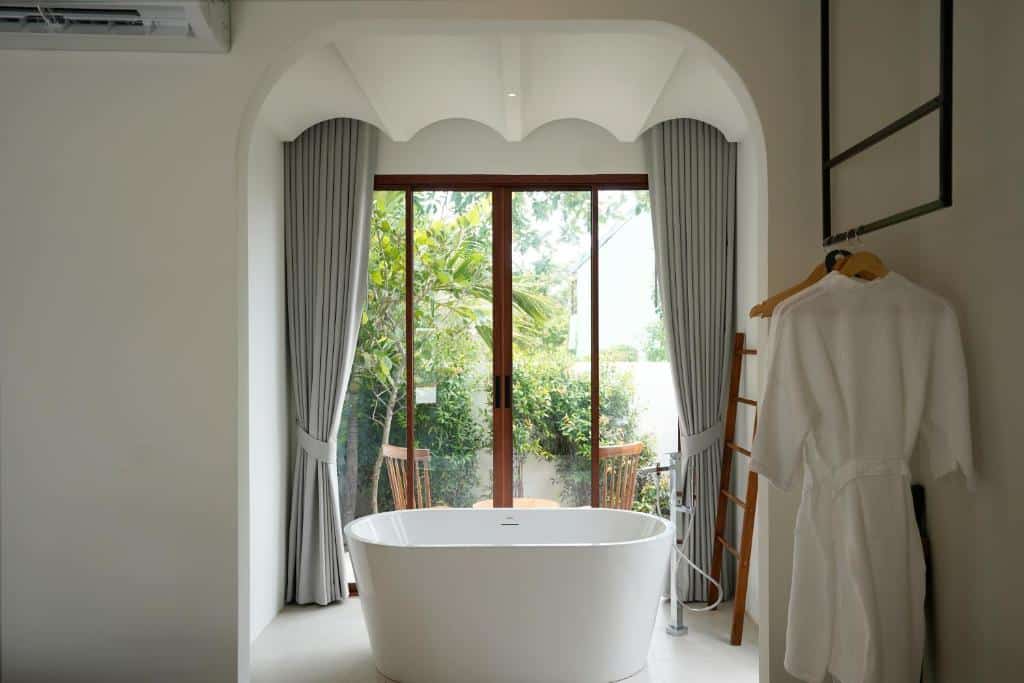 ห้องน้ำสไตล์โมเดิร์นพร้อมอ่างอาบน้ำสีขาวตั้งอยู่กลางห้อง, ผ้าม่านสีเทา, ประตูบานใหญ่กระจกใสที่มองเห็นต้นไม้สีเขียว, และผ้าเช็ดตัวสีขาวแขวนอยู่บนราว—เหมาะอย่างยิ่งหลังจากสำรวจ เกาะล้านที่พัก หรือผ่อนคลายที่ ที่พักเกาะล้านราคาถูก.