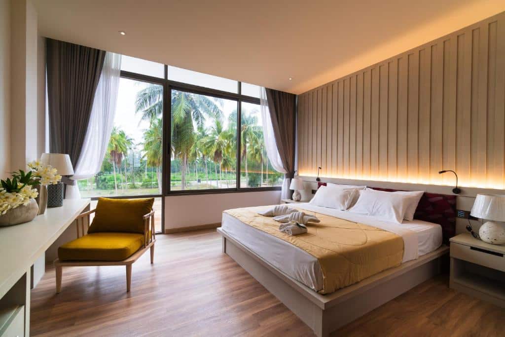 ห้องพักโรงแรมสมัยใหม่พร้อมเตียงขนาดใหญ่ เก้าอี้สีเหลือง โต๊ะทำงาน และหน้าต่างบานใหญ่จากพื้นจรดเพดานที่มองเห็นต้นปาล์มด้านนอก แสงไฟอบอุ่นและการตกแต่งแบบมินิมอลทำให้ที่พักติดทะเลประจวบแห่งนี้เหมาะสำหรับการพักผ่อนริมชายฝั่ง.