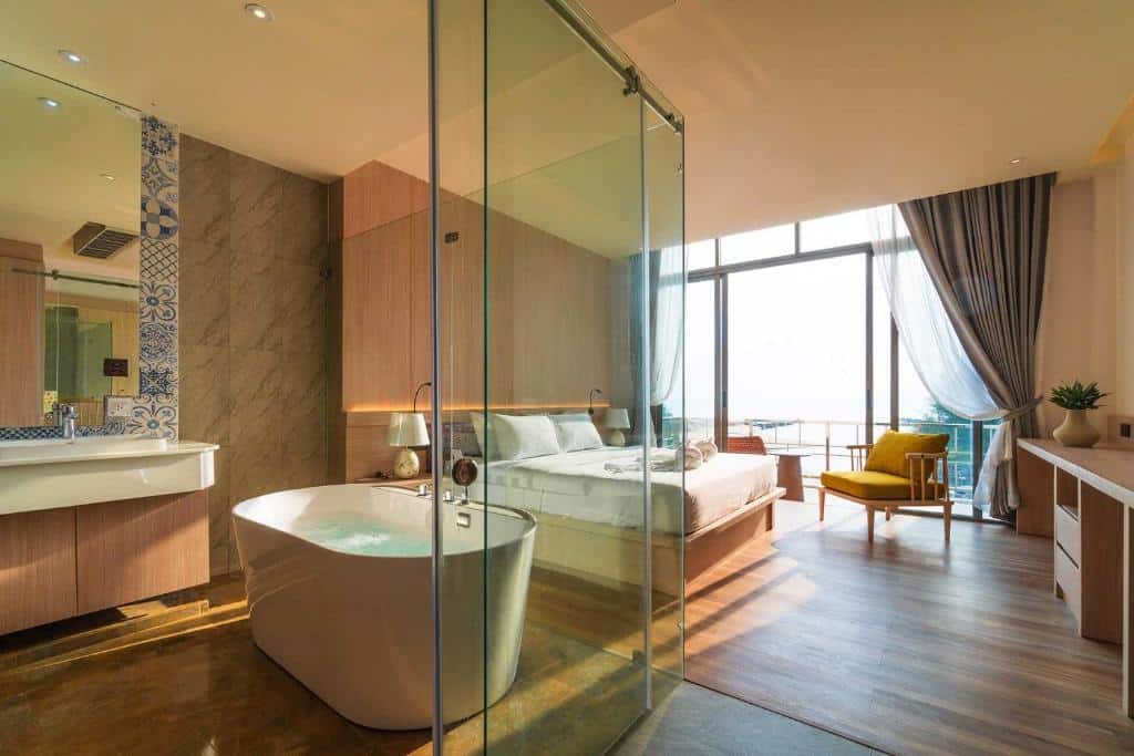 ห้องพักโรงแรมสมัยใหม่พร้อมห้องน้ำกระจก, อ่างอาบน้ำแบบลอยตัว, เตียง, เก้าอี้นวม, หน้าต่างบานใหญ่, และระเบียงพร้อมวิว—เหมาะสำหรับผู้ที่ต้องการที่พักประจวบติดทะเลที่มีสไตล์.