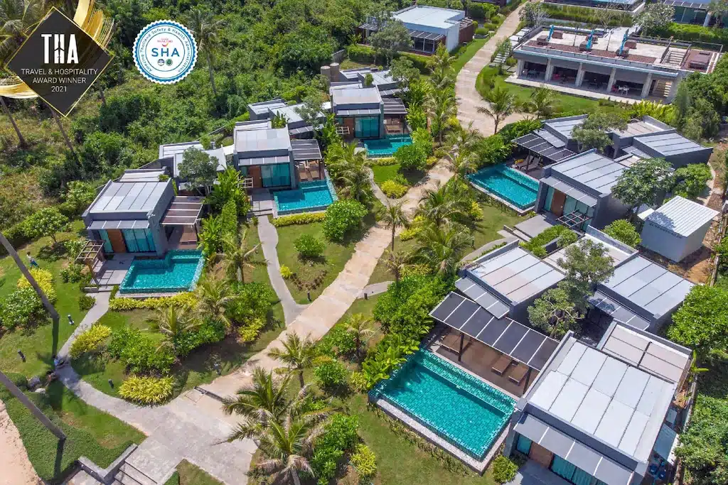 ภาพมุมสูงของรีสอร์ทที่พักประจวบคีรีขันธ์ พร้อมวิลล่าสมัยใหม่หลายหลัง แต่ละหลังมีสระว่ายน้ำส่วนตัว ล้อมรอบด้วยพื้นที่สีเขียวและทางเดิน; มีโลโก้รางวัลสองรางวัลปรากฏอยู่ตรงมุมบนซ้าย.