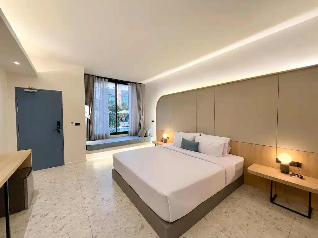 ห้องพักโรงแรมสมัยใหม่ที่ โรงแรมประจวบ พร้อมเตียงขนาดใหญ่ โต๊ะข้างเตียงพร้อมโคมไฟ ที่นั่งริมหน้าต่าง โต๊ะทำงาน และประตูสีฟ้า ตกแต่งด้วยโทนสีเรียบและของตกแต่งน้อยชิ้นเพื่อการพักผ่อนที่ผ่อนคลายในที่พักประจวบ.