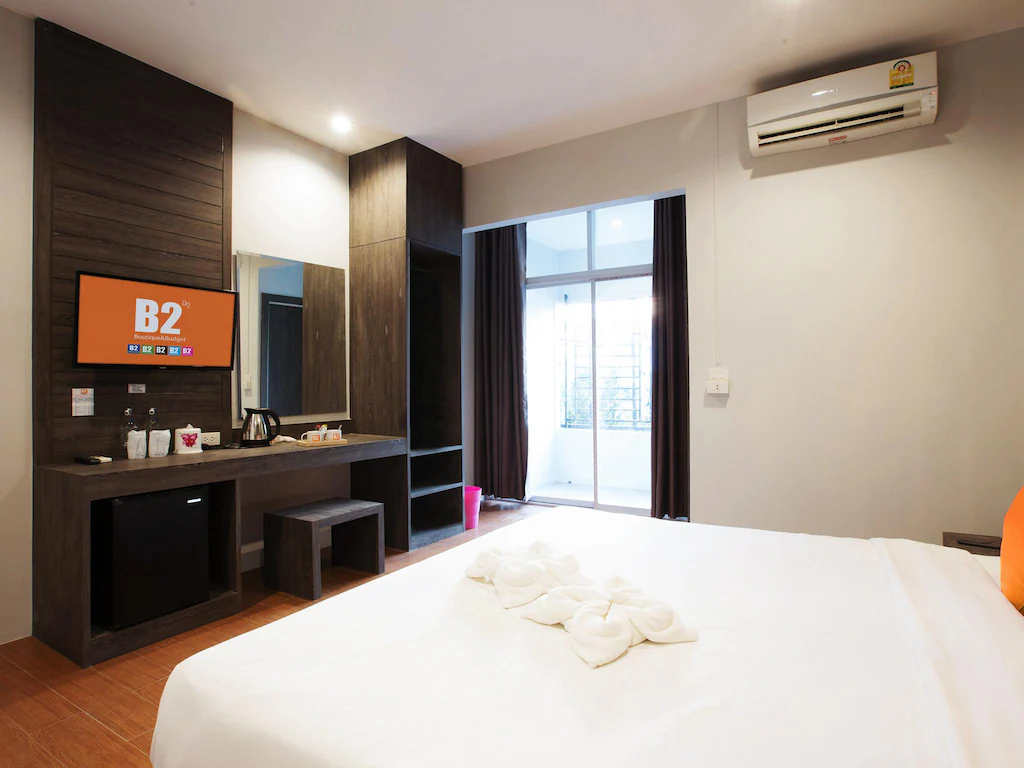 ห้องพักโรงแรมทันสมัยพร้อมเตียงคู่, โทรทัศน์จอแบน, โต๊ะทำงาน, ตู้เย็นขนาดเล็ก, ชุดชงชา, และเครื่องปรับอากาศ; ประตูระเบียงที่เปิดรับแสงธรรมชาติ—เหมาะสำหรับการผ่อนคลายหลังจากสำรวจสถานที่ท่องเที่ยวในบุรีรัมย์.