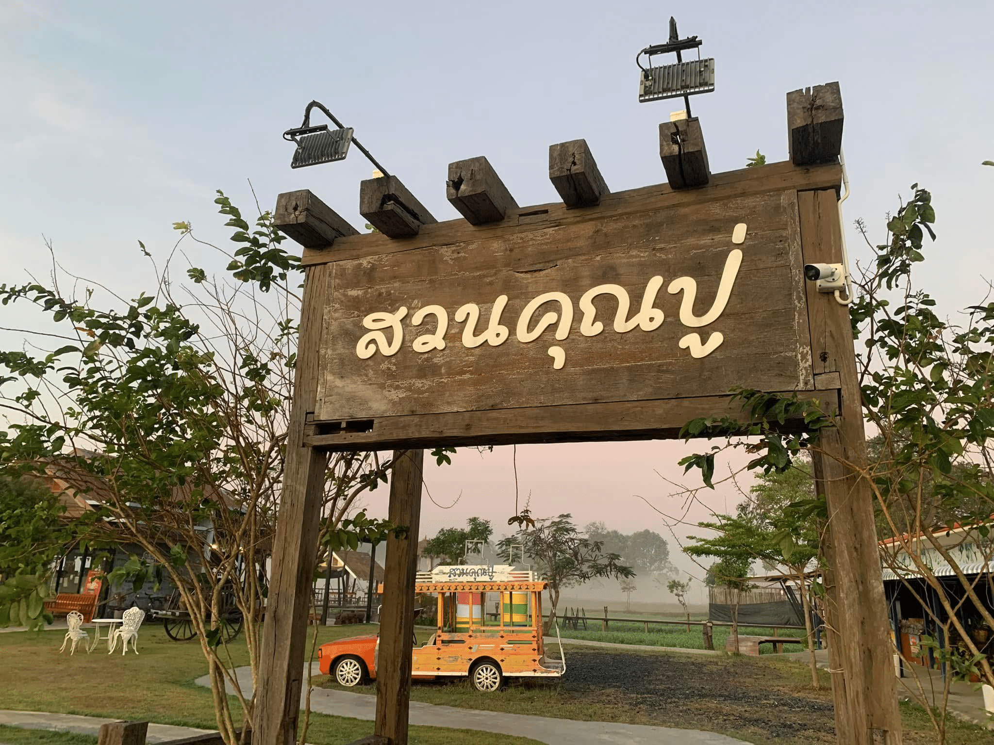 ป้ายไม้ที่มีตัวอักษรไทยตั้งอยู่ที่ทางเข้าของพื้นที่สวนสาธารณะใน ที่เที่ยวบุรีรัมย์ ล้อมรอบด้วยต้นไม้ ที่นั่ง และรถของเล่นในพื้นหลัง.