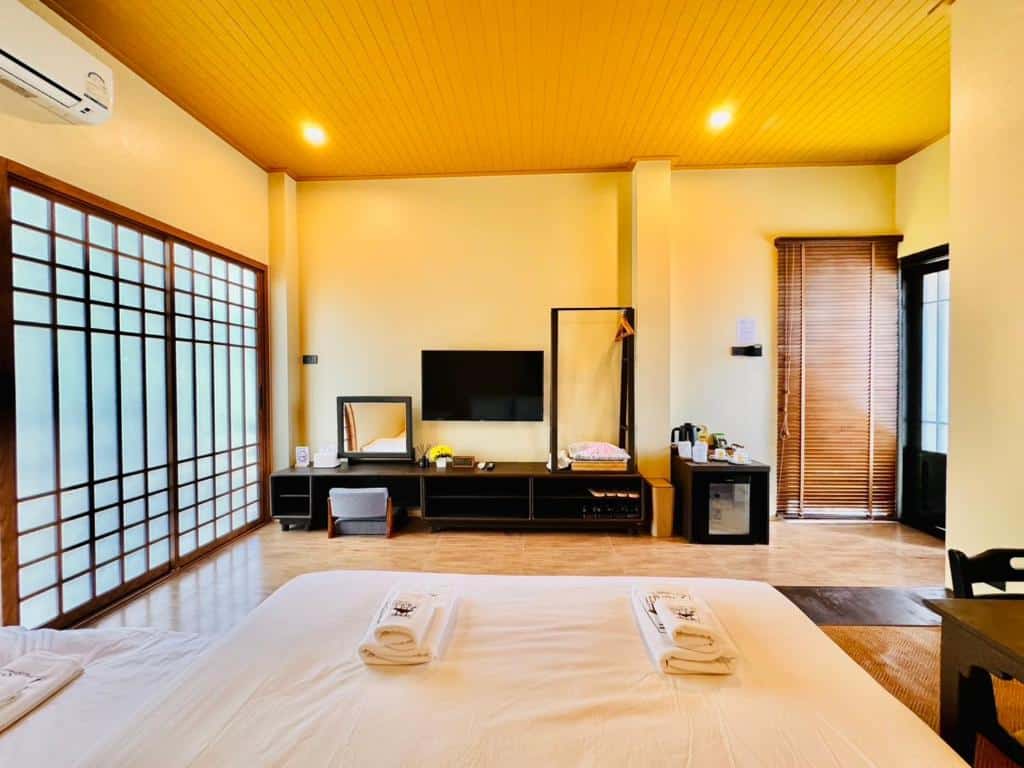 ห้องนอนสมัยใหม่พร้อมเตี้ย ผ้าเช็ดตัวพับเรียบร้อย โทรทัศน์บนคอนโซล ตู้เสื้อผ้า ตู้เย็นขนาดเล็ก และประตูบานเลื่อนสไตล์ญี่ปุ่น แสงไฟอบอุ่นและเพดานไม้ให้ความสบายหลังจากสำรวจสถานที่ท่องเที่ยวในบุรีรัมย์.
