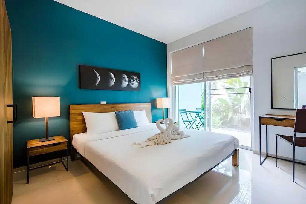 ห้องนอนสมัยใหม่ที่โรงแรมประจวบ ตกแต่งด้วยผนังสีเขียวอมฟ้าเข้ม เฟอร์นิเจอร์ไม้ เตียงขนาดใหญ่พร้อมผ้าขนหนูพับเป็นรูปหงส์ งานศิลปะรูปพระจันทร์เสี้ยว และหน้าต่างที่มองเห็นลานระเบียงเล็กๆ ที่เต็มไปด้วยต้นไม้สีเขียว.