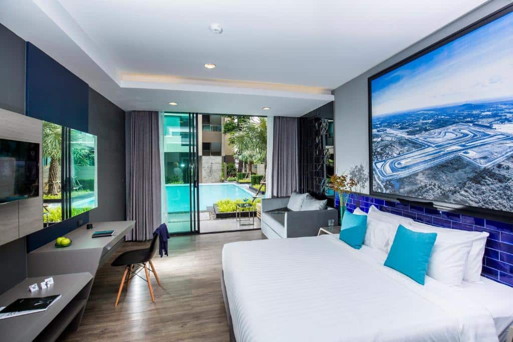 ห้องพักโรงแรมสมัยใหม่พร้อมเตียงขนาดใหญ่ โต๊ะทำงาน โซฟา ทีวีติดผนัง และระเบียงที่มองเห็นสระว่ายน้ำและต้นปาล์ม—เหมาะสำหรับการพักผ่อนหลังจากสำรวจบุรีรัมย์ที่เที่ยว หรือสถานที่ท่องเที่ยวบุรีรัมย์ที่น่าสนใจอื่นๆ ในบริเวณใกล้เคียง.