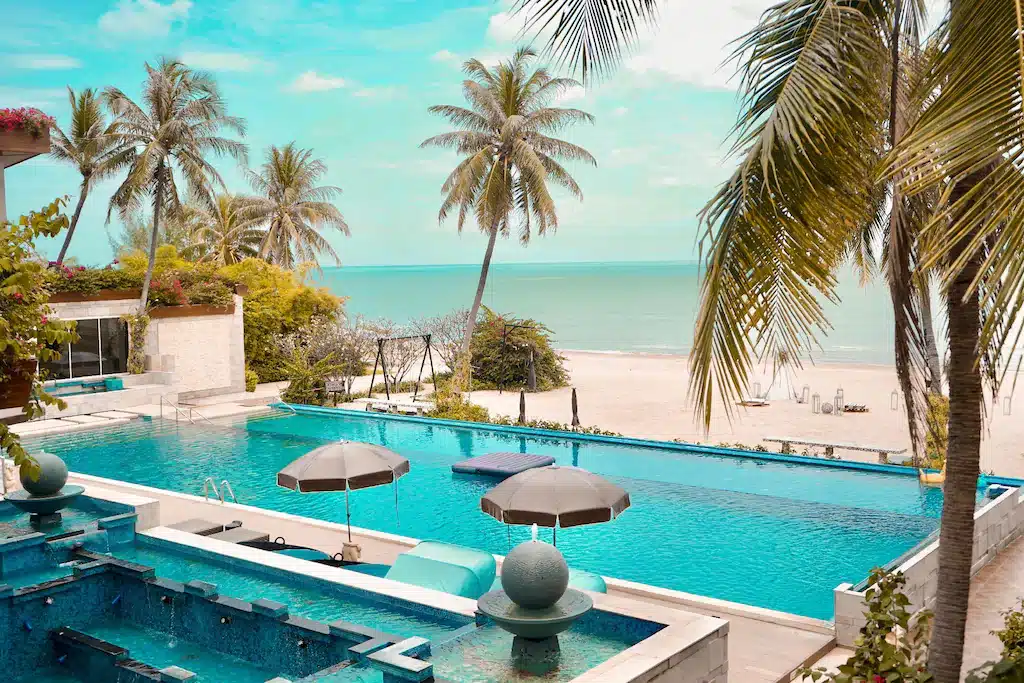 เพลิดเพลินกับการพักผ่อนที่สระว่ายน้ำริมชายหาดพร้อมเก้าอี้อาบแดด ร่ม ต้นปาล์ม และชายหาดทรายที่ทอดยาวสู่ทะเล—เหมาะสำหรับการเข้าพักครั้งต่อไปของคุณที่ โรงแรมประจวบ ท่ามกลางท้องฟ้าที่มีเมฆบางส่วน.