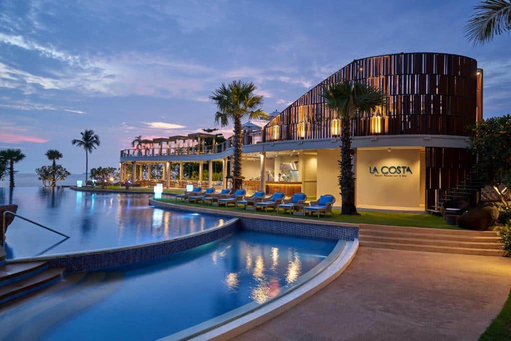รีสอร์ทริมชายหาดสมัยใหม่พร้อมสระว่ายน้ำโค้ง เก้าอี้อาบแดด ต้นปาล์ม และร้านอาหาร LA COSTA ในยามพระอาทิตย์ตกดิน—ตัวเลือกที่สมบูรณ์แบบสำหรับ ที่พักติดทะเลพัทยา หรือ โรงแรมติดทะเลพัทยา ของคุณ.
