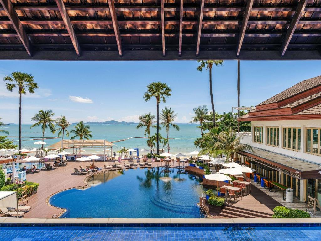 รีสอร์ทริมชายหาดในพัทยา มีสระว่ายน้ำล้อมรอบด้วยเก้าอี้อาบแดด ร่ม ต้นปาล์ม และวิวทะเลที่สวยงาม เหมาะสำหรับผู้ที่ต้องการโรงแรมพัทยาติดทะเลภายใต้ท้องฟ้าแจ่มใส.