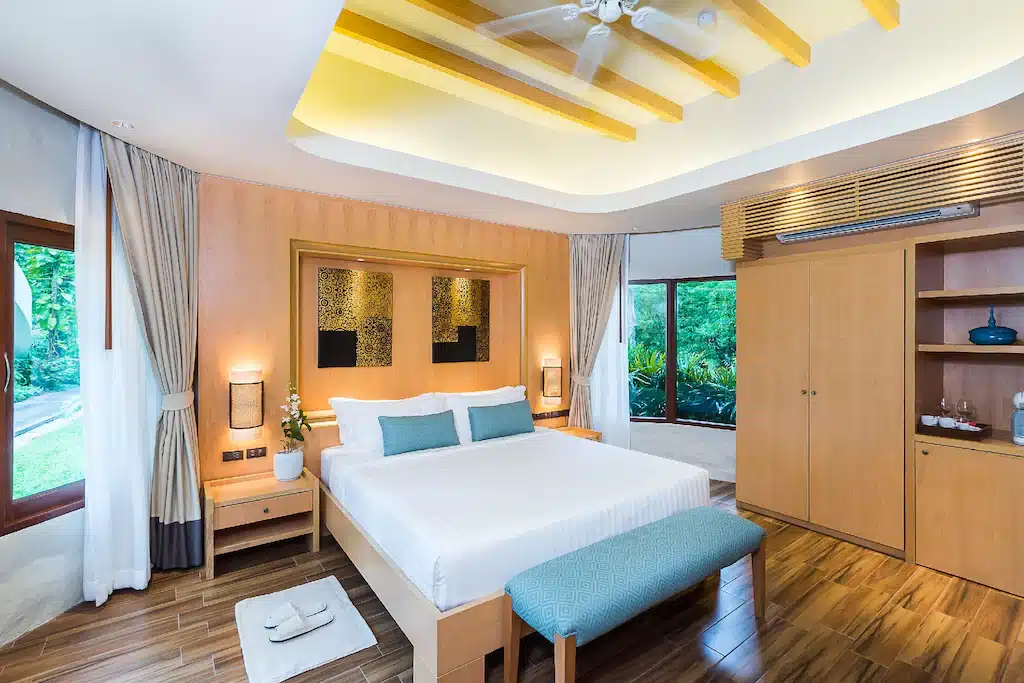 ห้องนอนสมัยใหม่พร้อมเตียงขนาดใหญ่ เฟอร์นิเจอร์ไม้ เพดานตกแต่งสวยงาม และหน้าต่างที่มองเห็นต้นไม้สีเขียวด้านนอก—เหมาะสำหรับการพักผ่อนในโรงแรมติดทะเลพัทยา พร้อมโต๊ะข้างเตียง ตู้เสื้อผ้า และม้านั่งที่ปลายเตียง.
