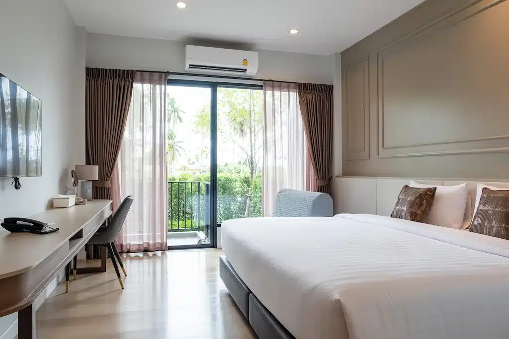ห้องพักโรงแรมประจวบสไตล์โมเดิร์น พร้อมเตียงขนาดใหญ่ โต๊ะทำงาน โทรทัศน์จอแบน และประตูบานเลื่อนกระจกที่เปิดออกสู่ระเบียงพร้อมวิวทิวทัศน์ภายนอกที่สวยงาม.