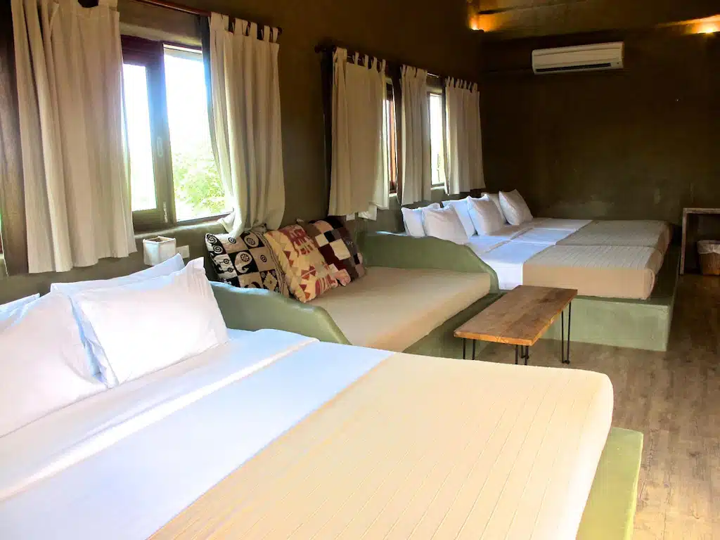 ห้องพักกว้างขวางที่รีสอร์ทปราณบุรี พร้อมเตียงใหญ่สามเตียง ผ้าปูที่นอนสีขาว โซฟา หมอนประดับ โต๊ะไม้ ผ้าม่านบางเบา และเครื่องปรับอากาศติดผนังเพื่อความสะดวกสบายของคุณ.