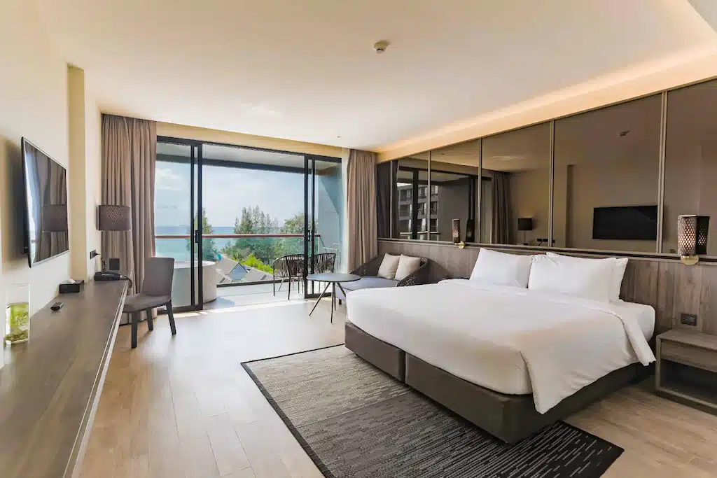 ห้องพักโรงแรมสมัยใหม่พร้อมเตียงขนาดใหญ่ โซฟา โต๊ะทำงาน โทรทัศน์จอแบน และระเบียงพร้อมเก้าอี้ที่สามารถมองเห็นวิวทะเลผ่านหน้าต่างบานใหญ่จากพื้นจรดเพดาน ที่ โรงแรมพัทยาติดทะเล สำหรับการพักผ่อนริมทะเลอย่างผ่อนคลาย.