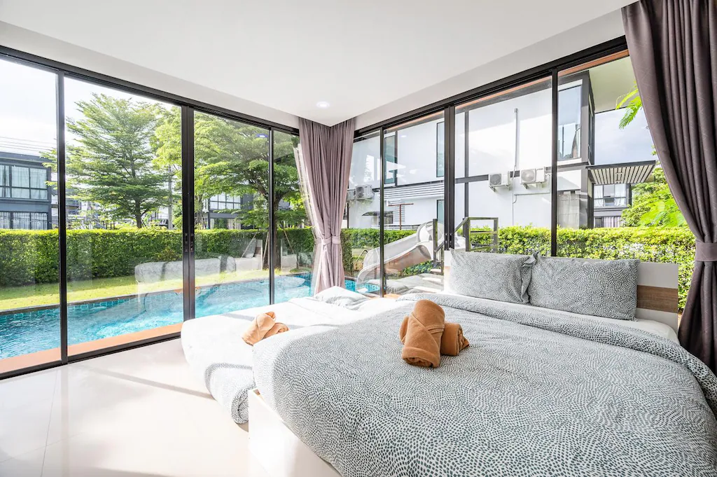 ห้องนอนสไตล์โมเดิร์นพร้อมหน้าต่างกระจกบานใหญ่ที่มองเห็นสระว่ายน้ำและสวนสวย มีเตียงที่จัดไว้อย่างเรียบร้อยพร้อมผ้าปูที่นอนลายสวย—เหมาะสำหรับการพักผ่อนในบ้านพักพูลวิลล่าพัทยาหรูหราสไตล์โมเดิร์นพร้อมวิวสระว่ายน้ำและสวนสวย.