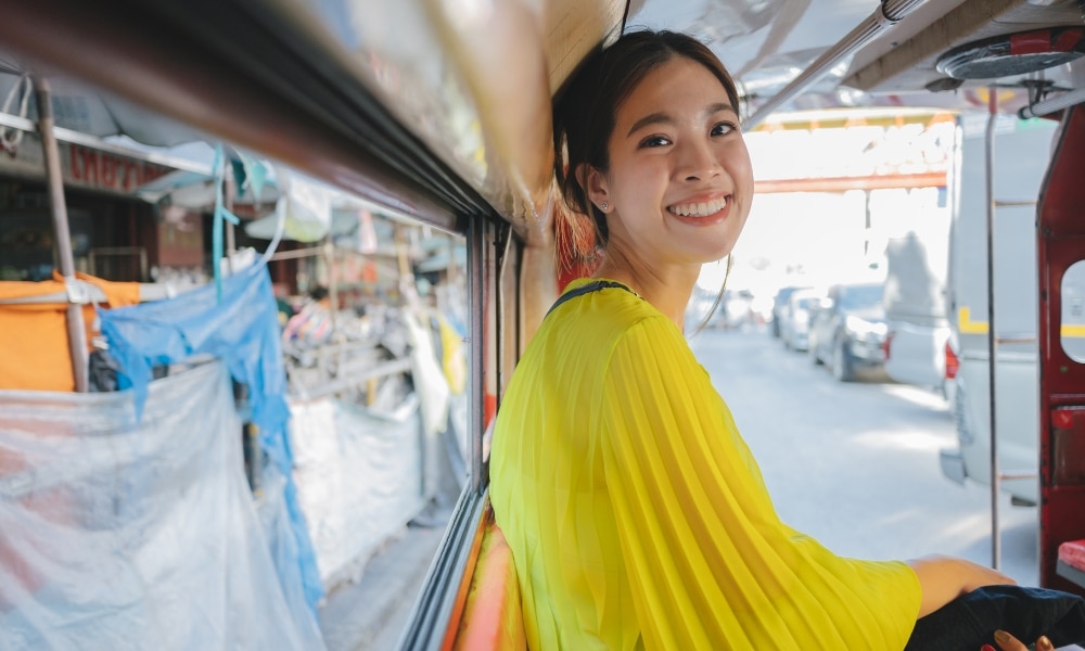 ผู้หญิงคนหนึ่งสวมเสื้อสีเหลืองสดใส นั่งอยู่ในรถ ยิ้มให้กล้อง พื้นหลังเป็นภาพถนนที่มีรถยนต์และแผงขายของในตลาด สร้างบรรยากาศที่มีชีวิตชีวาใกล้ที่พักธรรมชาติเชียงใหม่.