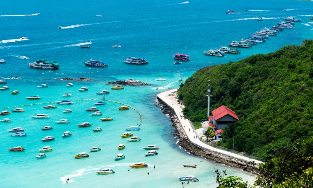 ภาพชายฝั่งที่มีเรือหลากสีสันมากมายในน้ำทะเลใสสีฟ้า ไฟสัญญาณที่มีหลังคาสีแดง และถนนคดเคี้ยวใกล้ที่พักติดทะเลพัทยาตามแนวเขาเขียวขจี.