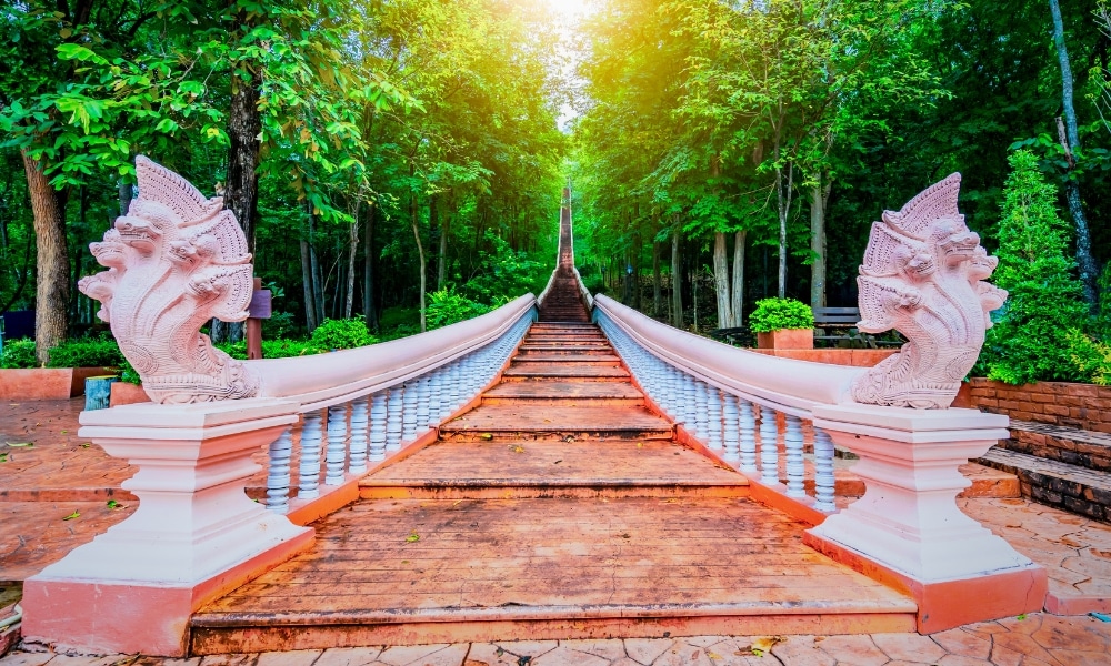 บันไดหินยาวที่มีราวบันไดสีขาวและรูปปั้นงูอยู่ทั้งสองข้าง นำทางขึ้นไปผ่านป่าเขียวชอุ่ม—สถานที่ที่น่าหลงใหลสำหรับผู้ที่ต้องการสำรวจ ที่เที่ยวบุรีรัมย์.