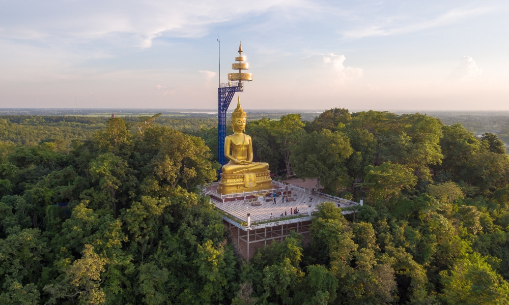 พระพุทธรูปทองคำขนาดใหญ่ตั้งอยู่บนแท่นล้อมรอบด้วยต้นไม้ในหนึ่งในสถานที่ท่องเที่ยวที่ต้องไปเยือนในบุรีรัมย์ โดยมีโครงสร้างตกแต่งสูงอยู่ข้างๆ และมีผู้คนมารวมตัวกันที่ฐานภายใต้ท้องฟ้าที่แจ่มใส.