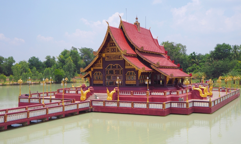 อาคารสไตล์ไทยดั้งเดิมที่มีหลังคาสีแดงประดับประดาอย่างวิจิตรตั้งอยู่บนแท่นเหนือสระน้ำ ล้อมรอบด้วยรูปปั้นมังกรสีทองและพืชพรรณเขียวชอุ่ม—สถานที่ท่องเที่ยวอันเป็นสัญลักษณ์ของบุรีรัมย์ที่เหมาะอย่างยิ่งสำหรับการดื่มด่ำกับวัฒนธรรมท้องถิ่น.