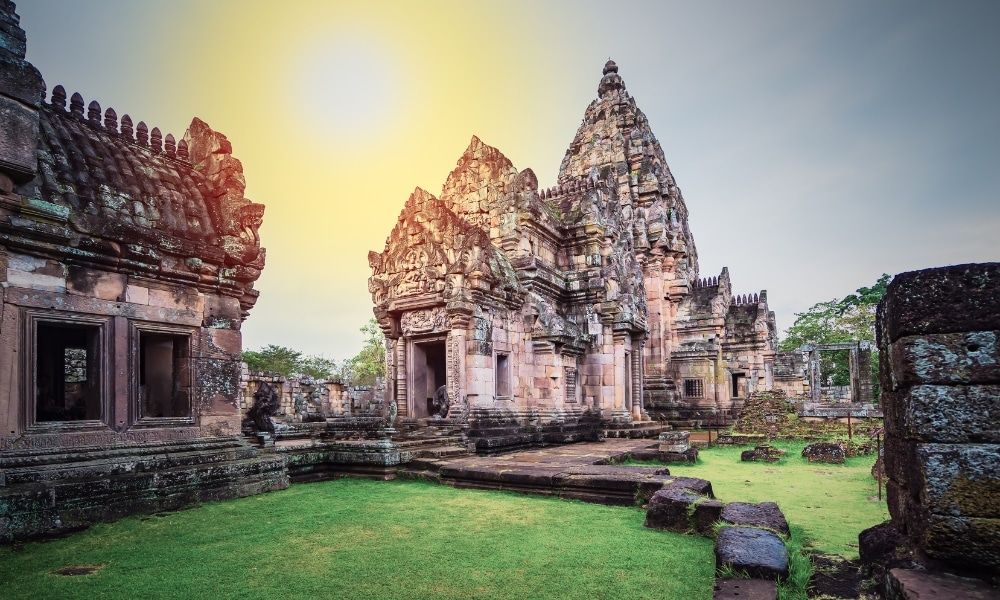 ซากปรักหักพังของวัดหินโบราณที่มีลวดลายแกะสลักอันซับซ้อนและหอคอยแหลม ตั้งอยู่ท่ามกลางสนามหญ้าสีเขียวภายใต้ท้องฟ้าสดใส—สถานที่ท่องเที่ยวบุรีรัมย์อันเป็นเอกลักษณ์ เหมาะสำหรับผู้ที่ต้องการสำรวจประวัติศาสตร์อันเป็นเอกลักษณ์และความงดงามของ เที่ยวบุรีรัมย์.