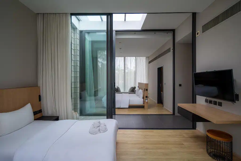 ห้องพักโรงแรมสมัยใหม่พร้อมเตียง, ทีวีติดผนัง, พื้นไม้, และผนังกระจกที่มองเห็นลานกลางแจ้ง—เหมาะสำหรับแขกที่ต้องการประสบการณ์ พูลวิลล่าพัทยา ที่มีสไตล์ใกล้ชายหาด.