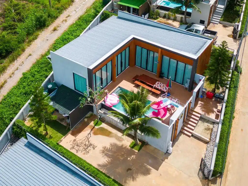 ภาพมุมสูงของบ้านพักตากอากาศแบบพูลวิลล่าชั้นเดียวสไตล์โมเดิร์นที่เขาค้อ พร้อมหน้าต่างบานใหญ่ สระว่ายน้ำส่วนตัวพร้อมของเล่นเป่าลม ต้นปาล์ม และถนนทางเข้าบ้านกว้างขวาง.