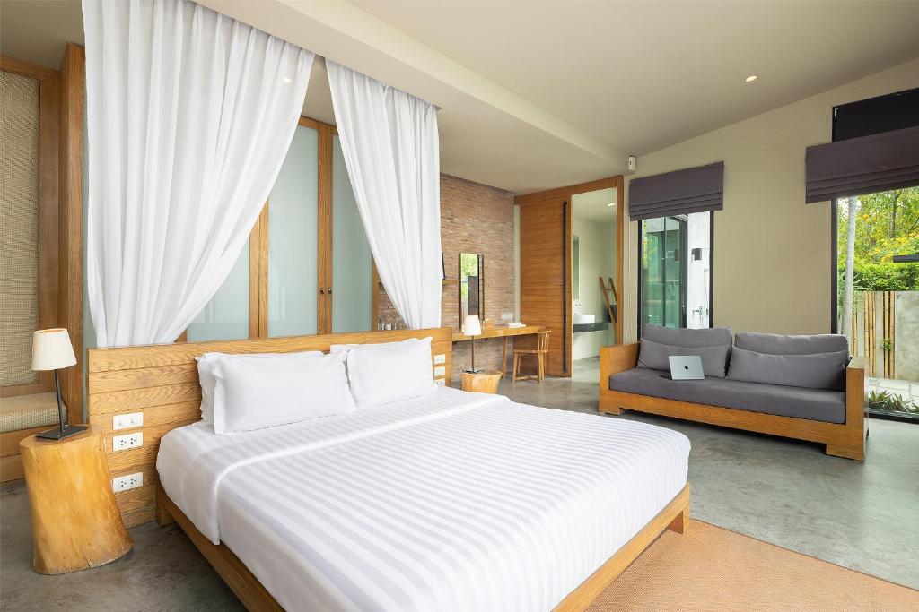 ห้องนอนสมัยใหม่พร้อมเตียงไม้ โครงเตียงไม้, ผ้าปูที่นอนลายทางสีขาว, โซฟาสีเทาพร้อมแลปท็อป, และหน้าต่างบานใหญ่จากพื้นจรดเพดานที่ส่องแสงสว่างตามธรรมชาติเข้ามา—เหมาะสำหรับการผ่อนคลายในบ้านพักพูลวิลล่าพัทยาของคุณหลังจากวันที่ไปเที่ยวชายหาดมา.