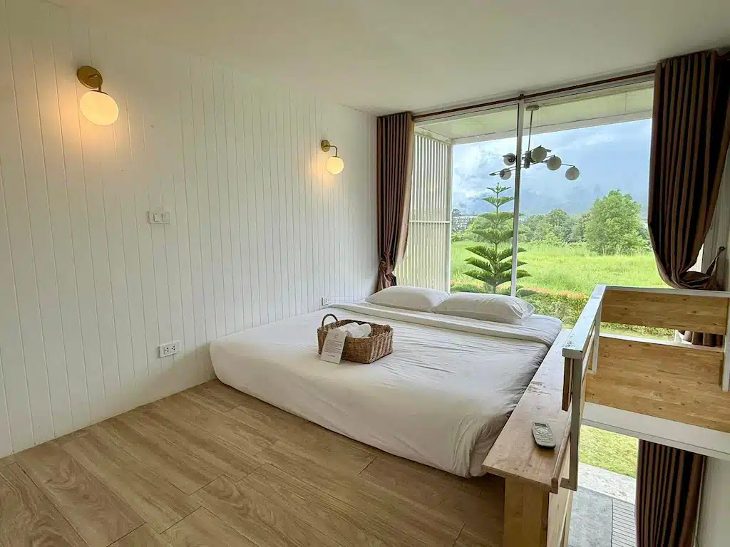ห้องนอนสไตล์มินิมอลพร้อมเตี้ยเตียงสีขาว ผ้าปูที่นอนสีขาว พื้นไม้ โคมไฟติดผนัง ตะกร้าหวาย และหน้าต่างบานใหญ่ที่มองเห็นทิวทัศน์สีเขียวและต้นไม้—เหมาะสำหรับการพักผ่อนอย่างเต็มที่ที่บ้านพักพูลวิลล่าเขาค้อ.