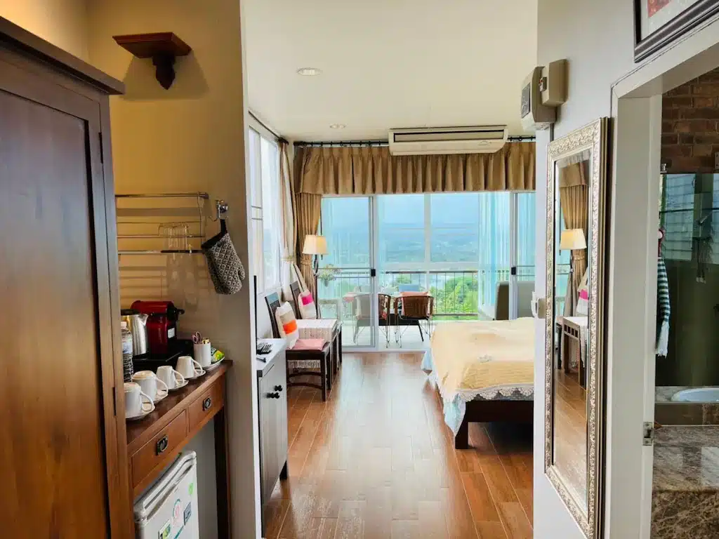 ห้องพักโรงแรมที่จัดเรียงอย่างเรียบร้อยพร้อมเตียง พื้นที่นั่งเล่น มุมกาแฟ และหน้าต่างบานใหญ่ที่เปิดออกสู่ระเบียงพร้อมวิวทิวทัศน์ที่งดงาม ณ พูลวิลล่าเขาค้อ.
