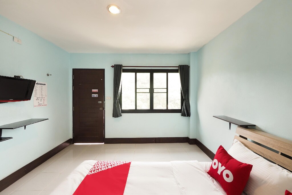 ห้องพักโรงแรมเรียบง่ายพร้อมผนังสีฟ้าอ่อน เตียงพร้อมผ้าปูที่นอนสีแดงและขาว หน้าต่างพร้อมผ้าม่านสีเข้ม โทรทัศน์ติดผนัง และประตู—เหมาะสำหรับการพักผ่อนอย่างสมบูรณ์แบบใกล้ที่พักเชียงใหม่ธรรมชาติ.