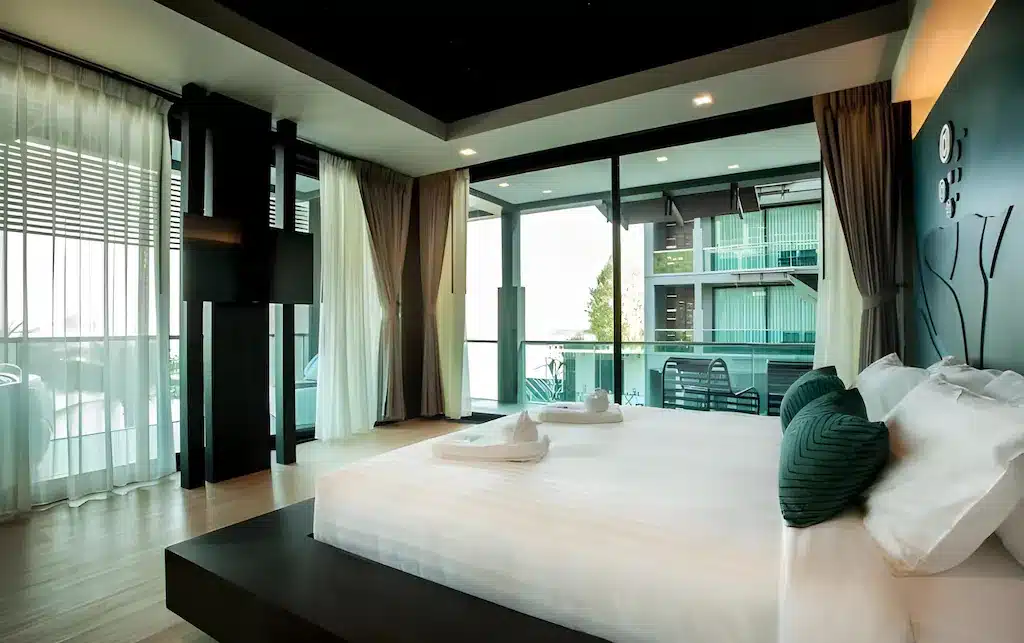 ห้องพักโรงแรมสมัยใหม่พร้อมเตียงขนาดใหญ่ หมอนอิงสีเขียวสดใส หน้าต่างบานใหญ่จากพื้นจรดเพดาน ผ้าม่านบางเบา และระเบียงพร้อมที่นั่งกลางแจ้ง—เหมาะสำหรับการพักผ่อนที่ ที่พักประจวบติดทะเล.