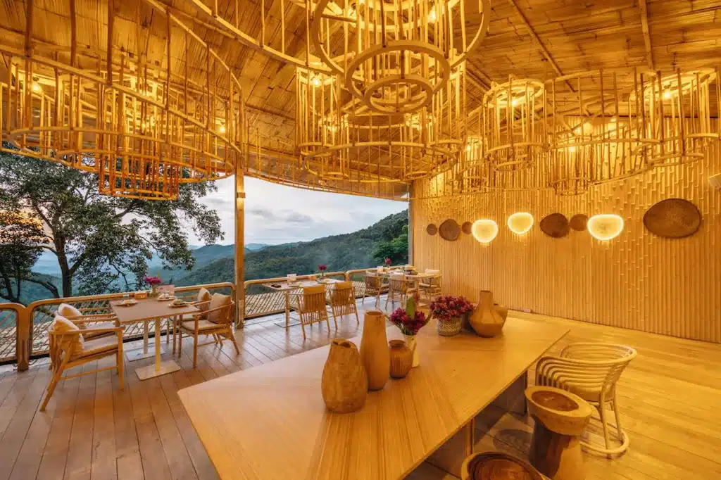 ร้านอาหารกลางแจ้งตกแต่งด้วยไม้ไผ่ เฟอร์นิเจอร์ไม้ และแสงไฟอบอุ่น มองเห็นวิวภูเขา เหมาะสำหรับผู้ที่มองหาที่พักเชียงใหม่ท่ามกลางธรรมชาติ ท่ามกลางต้นไม้และท้องฟ้าที่มีเมฆเป็นฉากหลัง.