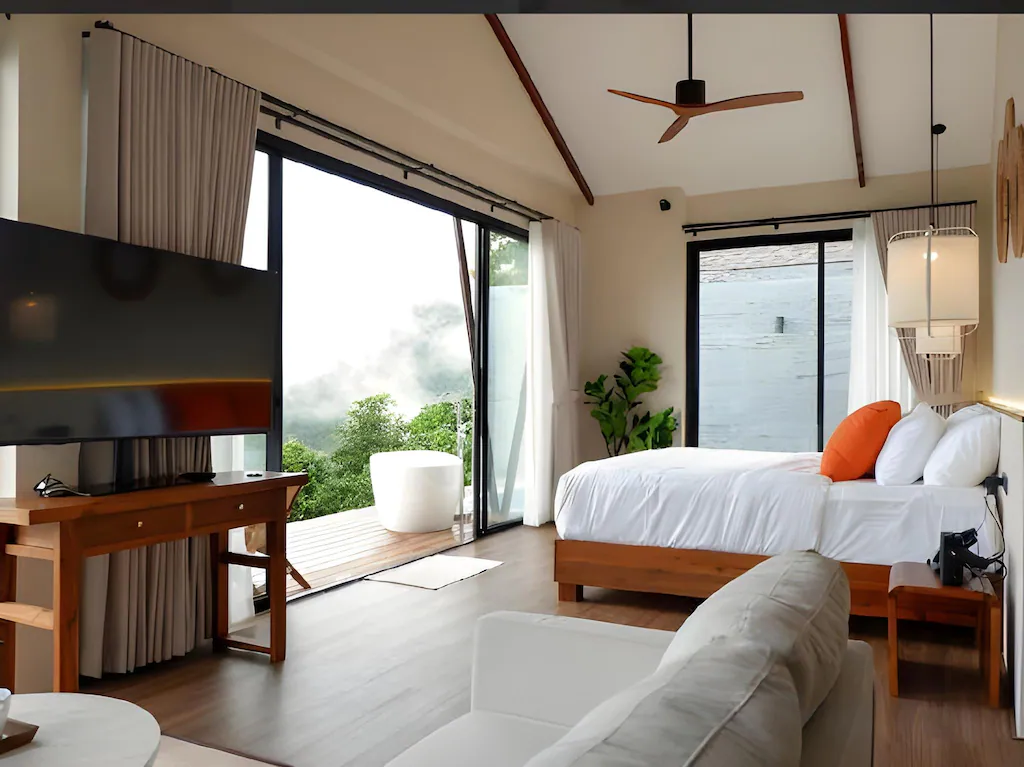 ห้องพักโรงแรมทันสมัยพร้อมหน้าต่างบานใหญ่ เตียงสีขาว หมอนสีส้ม ทีวี เฟอร์นิเจอร์ไม้ และพัดลมเพดาน เพลิดเพลินกับวิวธรรมชาติสีเขียวและอ่างอาบน้ำกลางแจ้งบนระเบียง—เหมาะสำหรับการพักผ่อน ที่พักเชียงใหม่ธรรมชาติ.