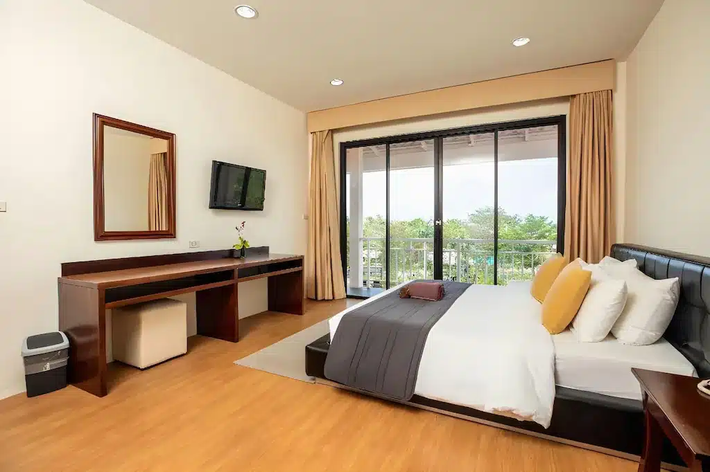 ห้องพักโรงแรมสมัยใหม่พร้อมเตียงขนาดใหญ่ หมอนหลายใบ โต๊ะพร้อมกระจก โทรทัศน์ติดผนัง และประตูกระจกบานเลื่อนไปยังระเบียงที่มีต้นไม้สีเขียว—เหมาะสำหรับผู้ที่ต้องการที่พักเกาะล้านติดทะเลในบรรยากาศผ่อนคลายและมีสไตล์.