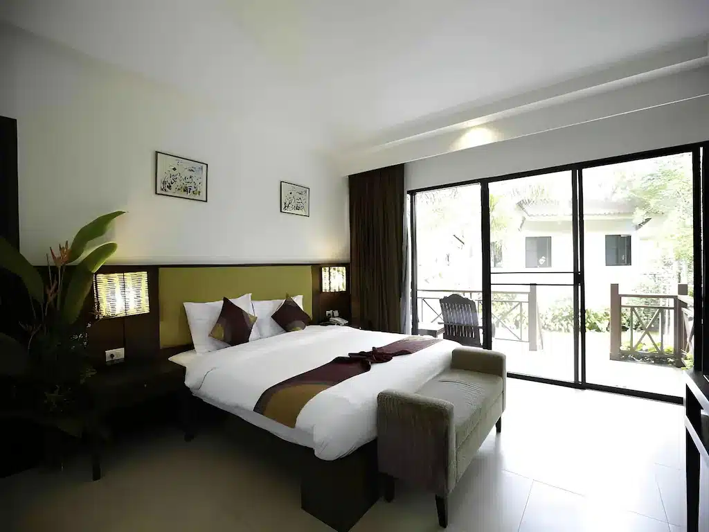 ห้องพักโรงแรมสมัยใหม่พร้อมเตียงคู่ที่จัดไว้อย่างเรียบร้อย โต๊ะข้างเตียง ต้นไม้ประดับ และประตูบานกระจกขนาดใหญ่ที่เปิดออกสู่ระเบียง—เหมาะสำหรับผู้ที่มองหาที่พักติดทะเล พร้อมวิวทะเลสวยงามและบรรยากาศผ่อนคลาย.