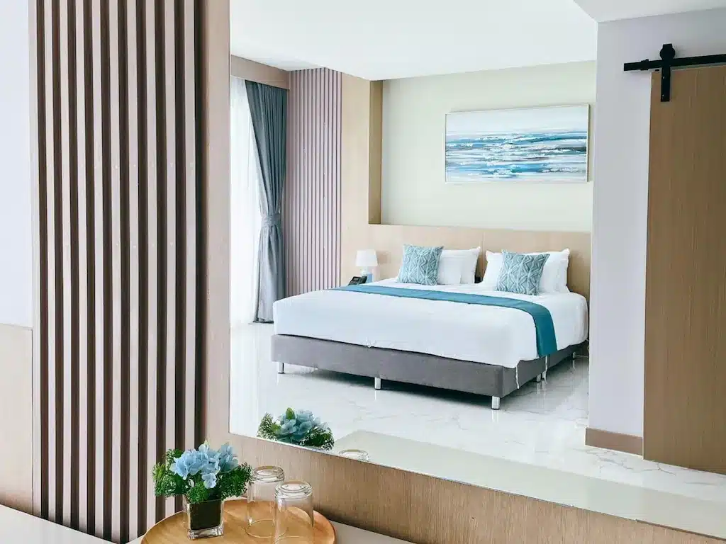 ห้องพักโรงแรมสมัยใหม่ที่ โรงแรมประจวบ พร้อมเตียงขนาดใหญ่ ตกแต่งด้วยสีน้ำเงิน โต๊ะข้างเตียง และงานศิลปะเหนือเตียง ประตูบานเลื่อนแบบโรงนาและภาพสะท้อนของโต๊ะเล็กที่มีขวดแก้วและดอกไม้ สร้างบรรยากาศที่พักติดทะเลประจวบที่มีสไตล์.