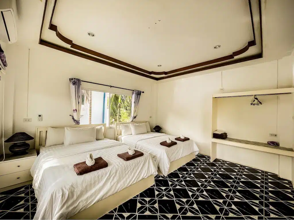 ห้องพักโรงแรมที่เรียบร้อยที่ ที่พักเกาะล้านติดทะเล พร้อมเตียงเดี่ยวสองเตียง ผ้าปูที่นอนสีขาว ผ้าเช็ดตัวพับเรียบร้อย หน้าต่างพร้อมผ้าม่าน ชั้นวางของเปิดโล่ง และพื้นกระเบื้องลายขาวดำ.
