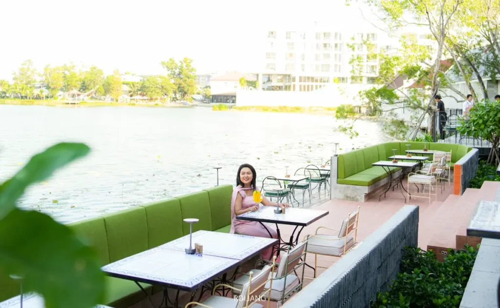 ผู้หญิงคนหนึ่งนั่งอยู่คนเดียวที่โต๊ะคาเฟ่กลางแจ้งใกล้ทะเลสาบที่ DAY & NIGHT เชิงทะเล พร้อมถือเครื่องดื่มไว้ในมือ โต๊ะอื่น ๆ และที่นั่งที่มีเบาะสีเขียวว่างเปล่า โดยมีต้นไม้และตึกอาคารให้เห็นอยู่เบื้องหลัง.